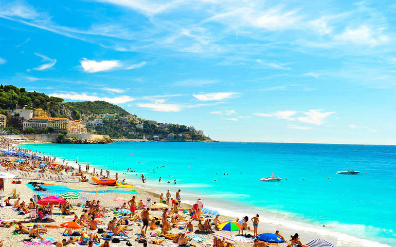 A popular Mediterranean beach filled with people and brightly coloured beach umbrellas with a clear blue sea and clear blue sky.