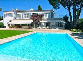Looking across the clear blue pool to the rear of a large white villa with a pristine green lawn on the left and to the right a table and chairs shaded by a large tree.