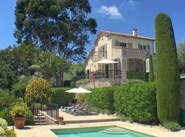 A pool in the foreground with the terrace and sun loungers beyond and a traditional 2-storey holiday villa in the background surroundd by mature gardens with hedges and a large palm.